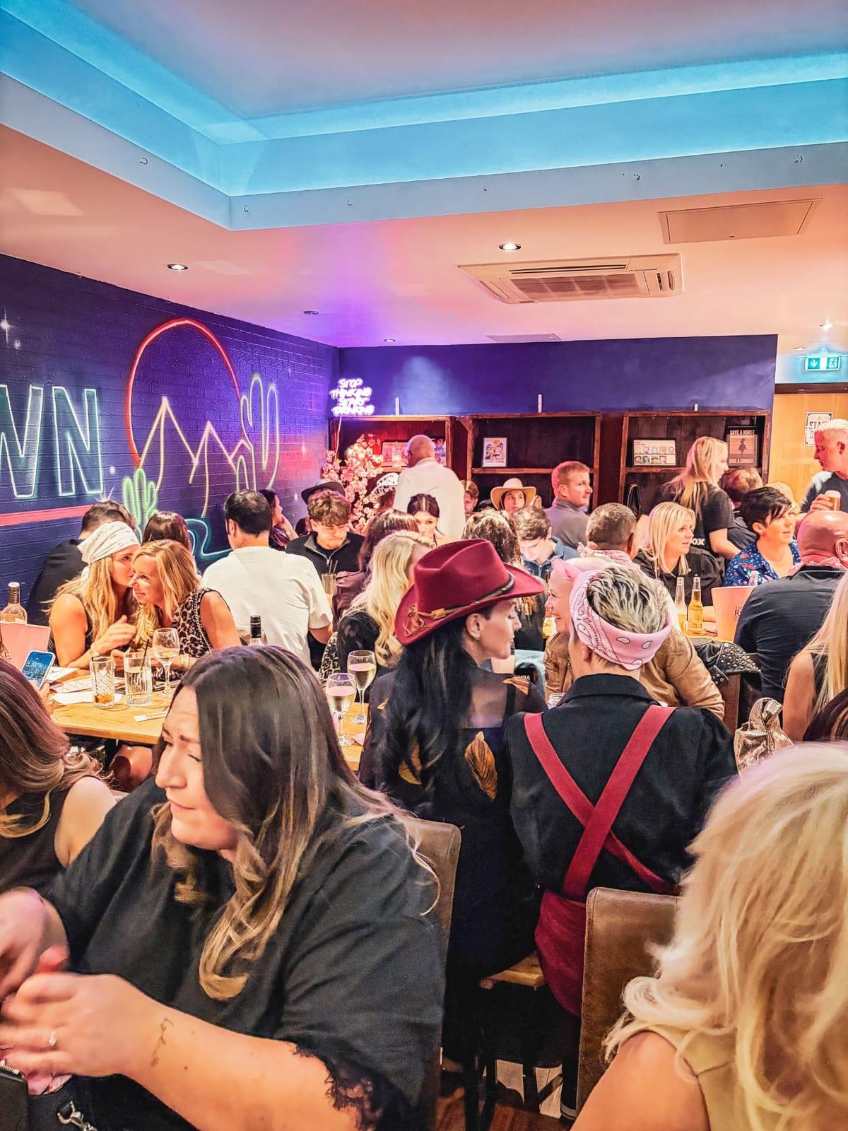 A packed HoeDown bar with cowboy hats, neon cactus artwork and full tables