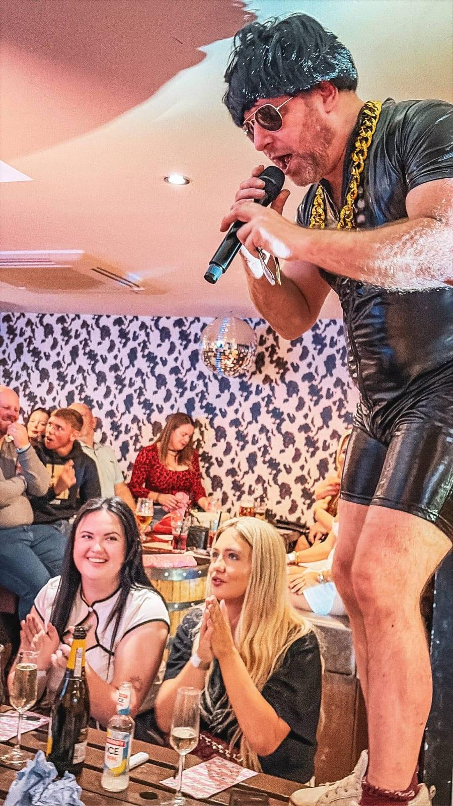 A performer in a wig and gold chain singing on a table to a cheering crowd at HoeDown
